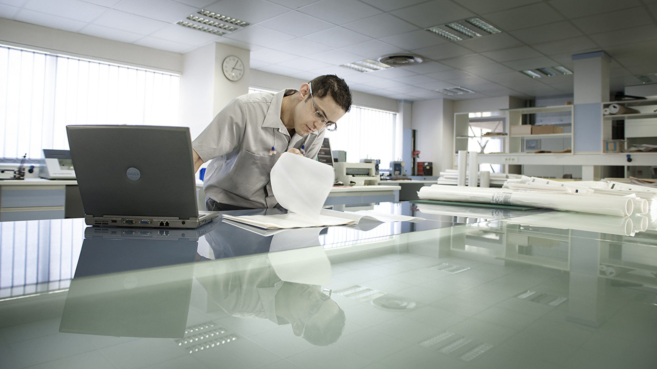 A laboratory technician at work, inspecting papers with a laptop computer in front of him in a big room. Corporate photography. Photographer Tuomas Harjumaaskola.