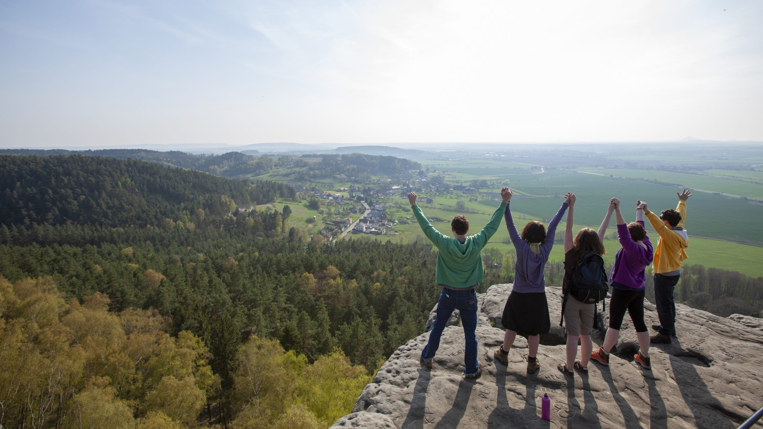 People holding raised hands at a teambuilding event on the top of a hill, with a view over the forest and fields. Photographer Tuomas Harjumaaskola.
