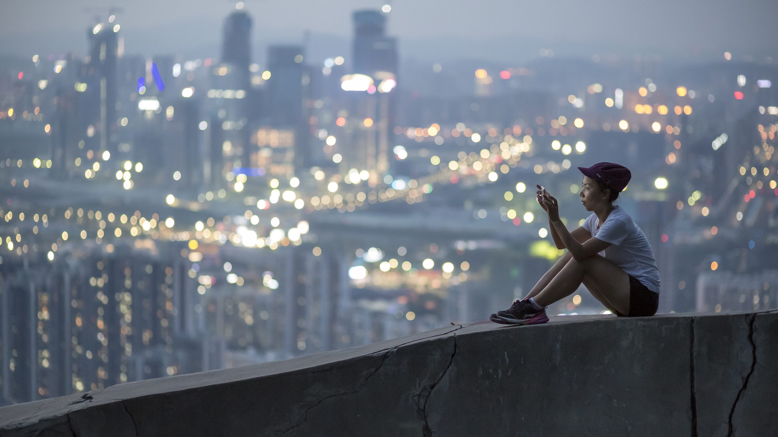 Young woman sitting on a hill in Shenzhen, China, the evening city lights behind her, holding a smartphone. Photographer Tuomas Harjumaaskola.