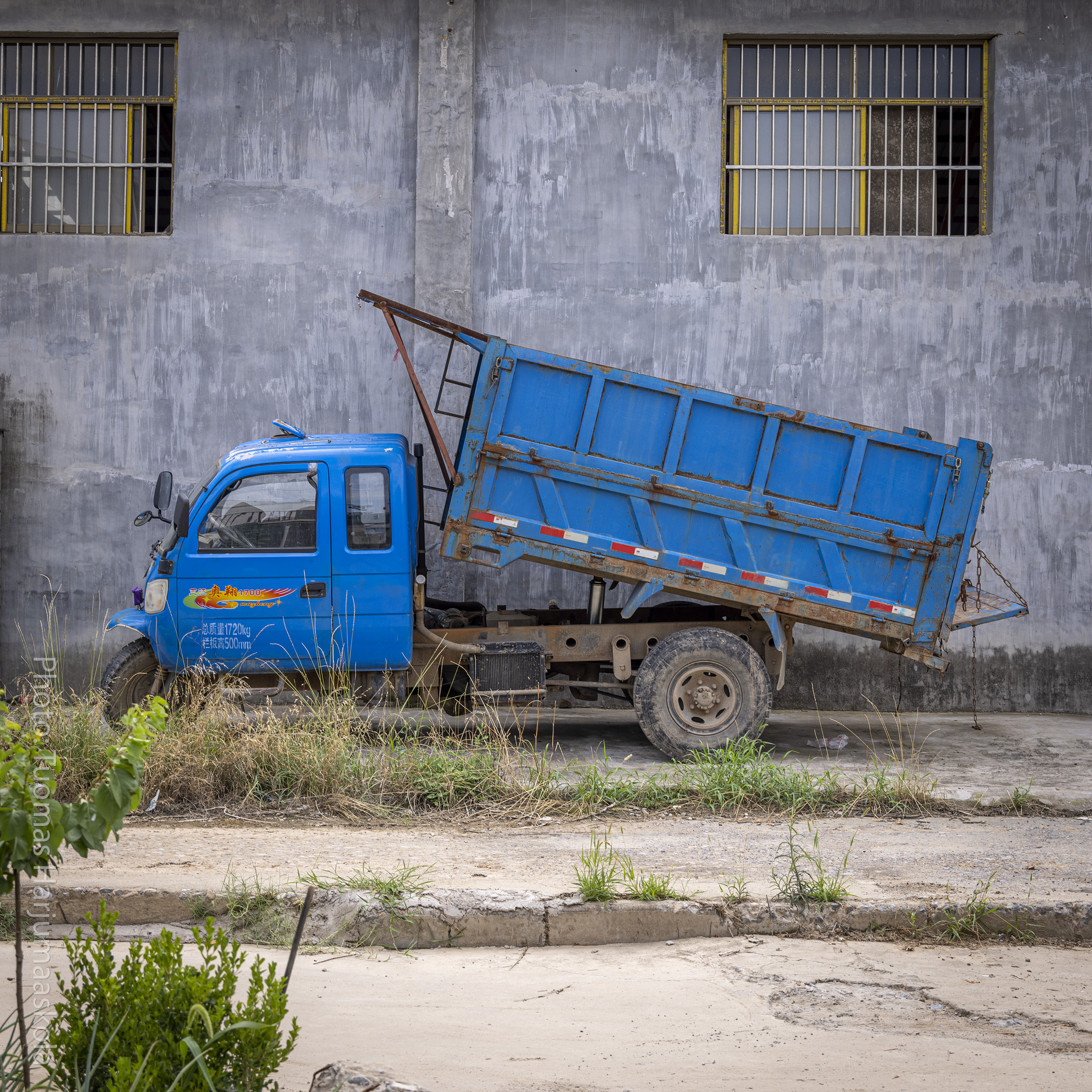 A blue Chinese tricycle truck, 3-wheeler truck with the bed lifted. Photographed against a concrete wall. Photographer Tuomas Harjumaaskola.
