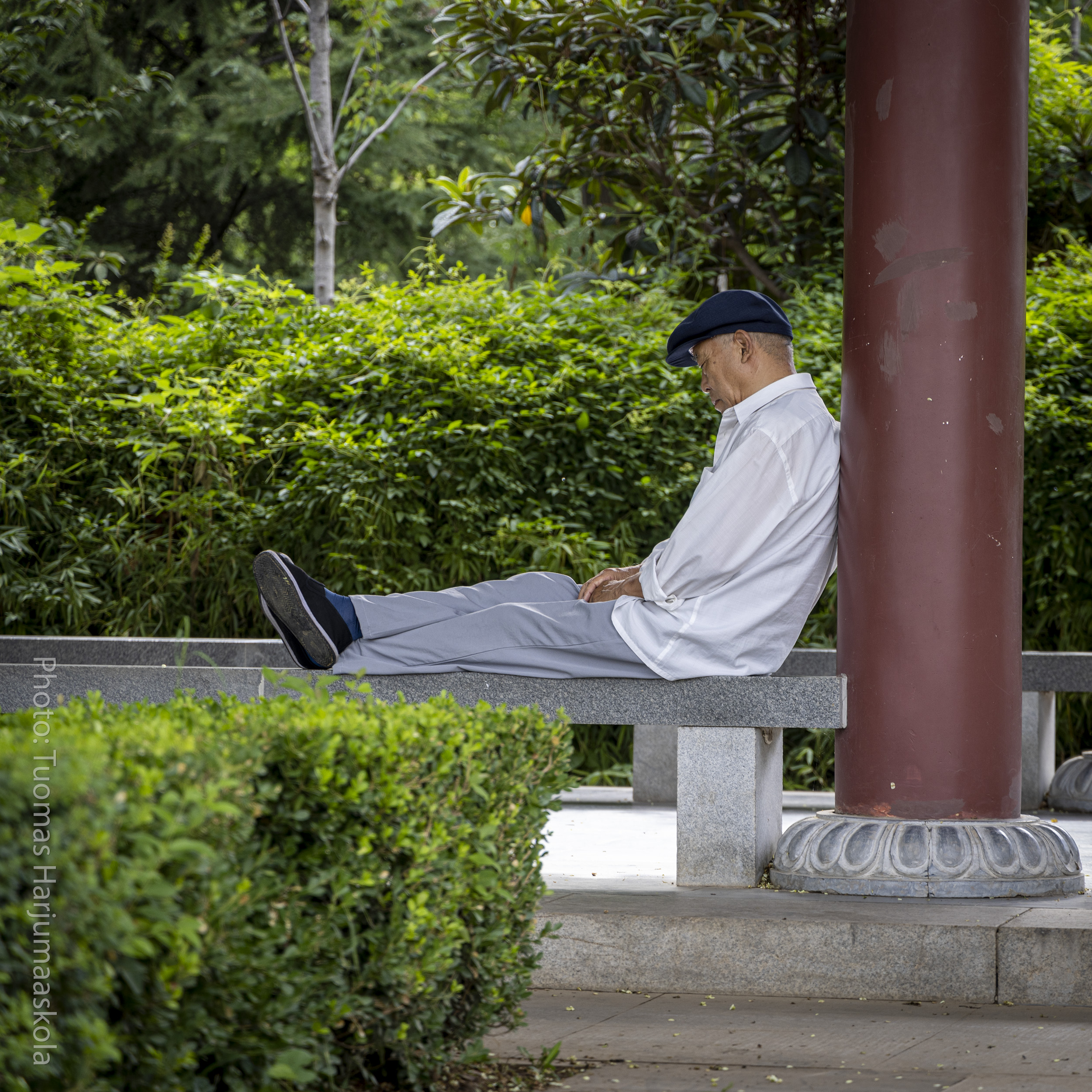 A Chinese senior man napping in the park, leaning against a column, wearing a hat. Photographer Tuomas Harjumaaskola.