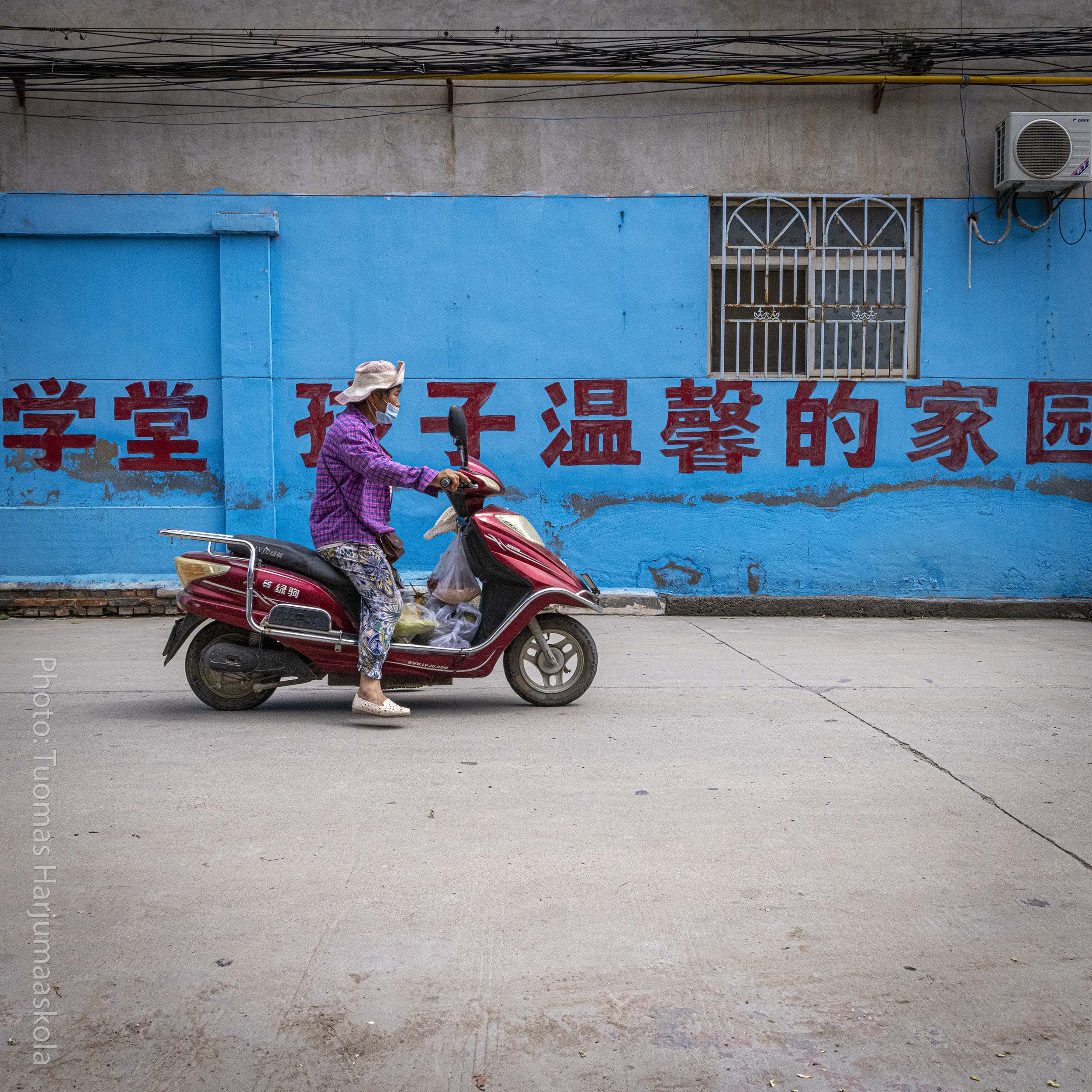 A woman passing by on a scooter, Chinese writing on a blue wall behind her. Photographer by Tuomas Harjumaaskola.